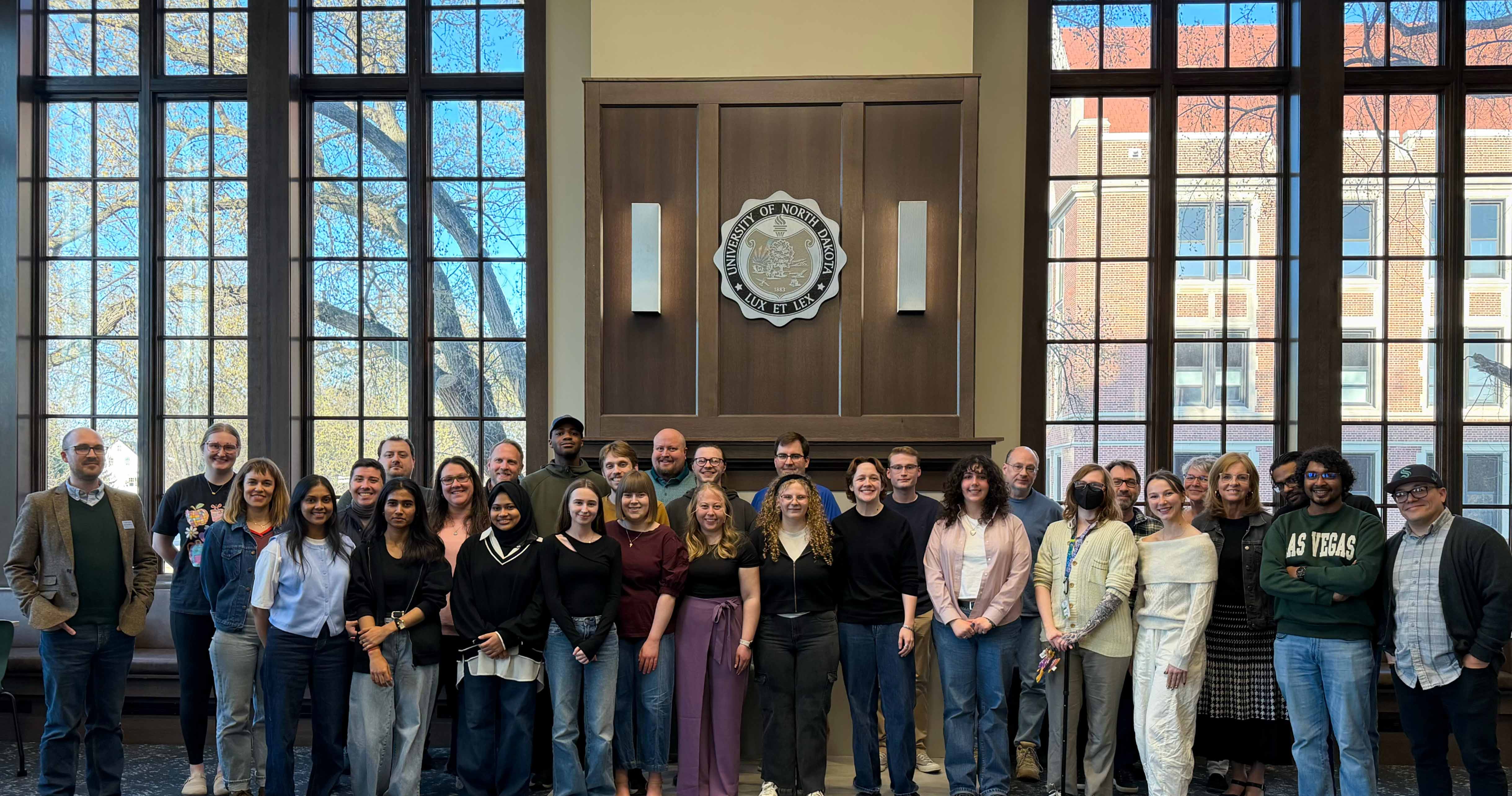 Group of students gathered in front of University of North Dakota seal on wood wall between windows. Housed in the library's reading and review room.