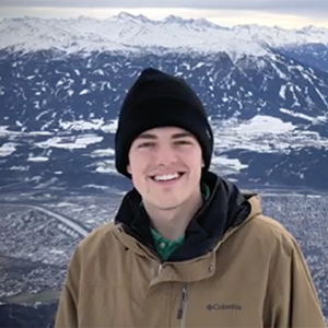 Student Austin Artz wears a hat and jacket smiling in front of a mountain range
