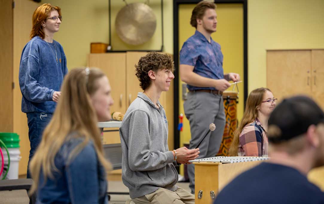 music educators sit in a circle on the floor playing guitar and hand percussion instruments