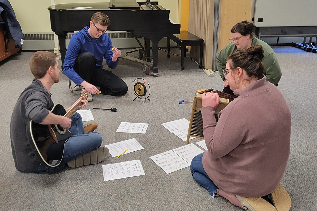 music educators sit in a circle on the floor playing guitar and hand percussion instruments