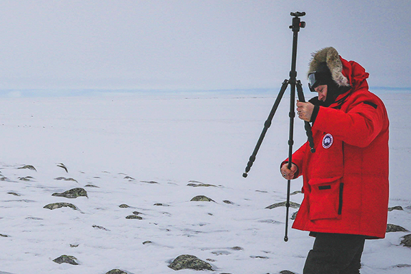 Snow and rocky landscape with Tim Pasch in an orange arctic coat setting up tripod equipment